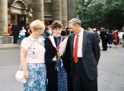 Michele wears a graduation gown in front of Convocation Hall with family